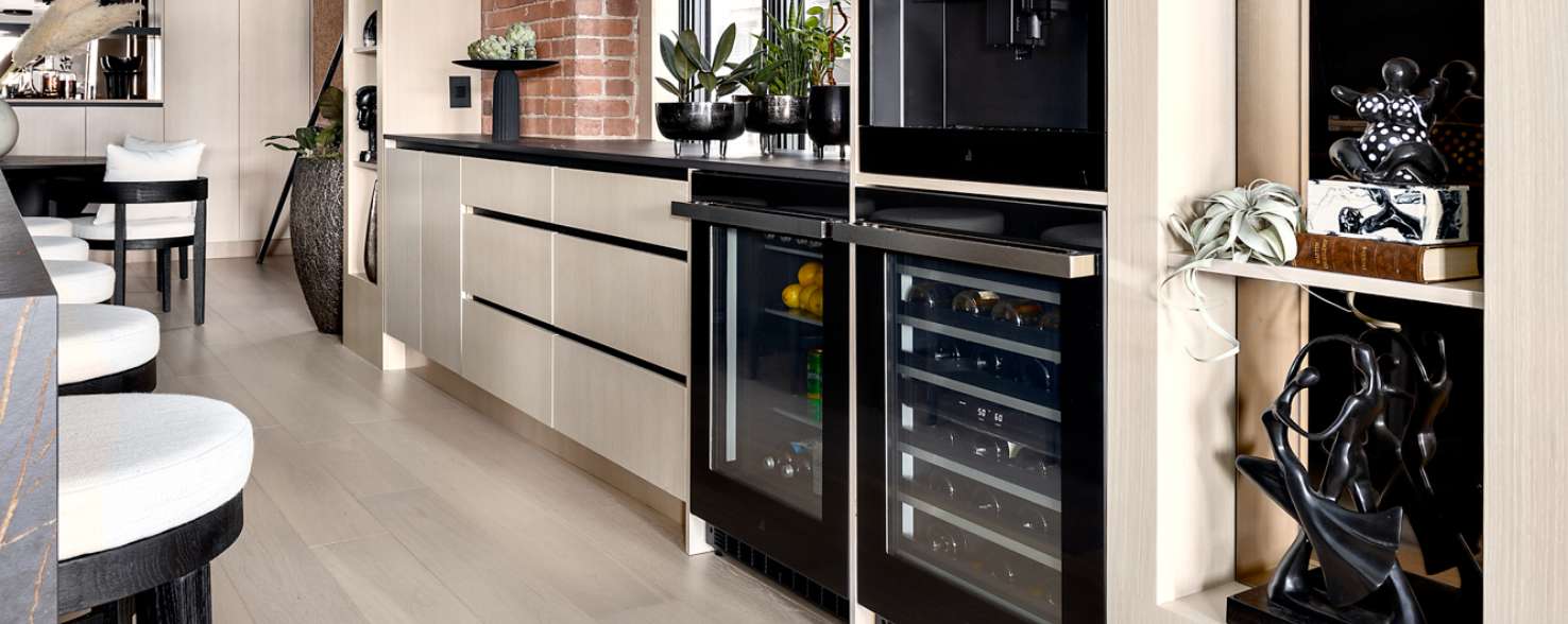 A kitchen with a JennAir Beverage Centre and Wine Cellar. Inside the beverage centre are lemons and various beverages. In the wine cellar are sideways bottles of wine. On the counter are various potted plants. Next to the wine cellar are shelves with statues on top. There's also a kitchen island with five black stools with white cushions. In the background is a black table and two white and black chairs.  