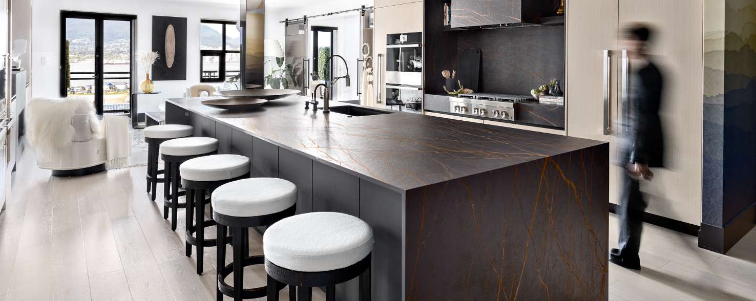  A blurry person stands in front of a panel-ready JennAir Double Refrigerator. Also in the kitchen is a large island with five black stools with white cushions and a JennAir Cooktop and Over-the-Range Vent Hood surrounded by a container of kitchen utensils, cutting boards and a bowl of fake produce. On top of the island are two decorative bowls. Behind the kitchen is a family room with a chair with a blanket over it, glass doors, windows, a potted plant, standing lamp and a vase. 