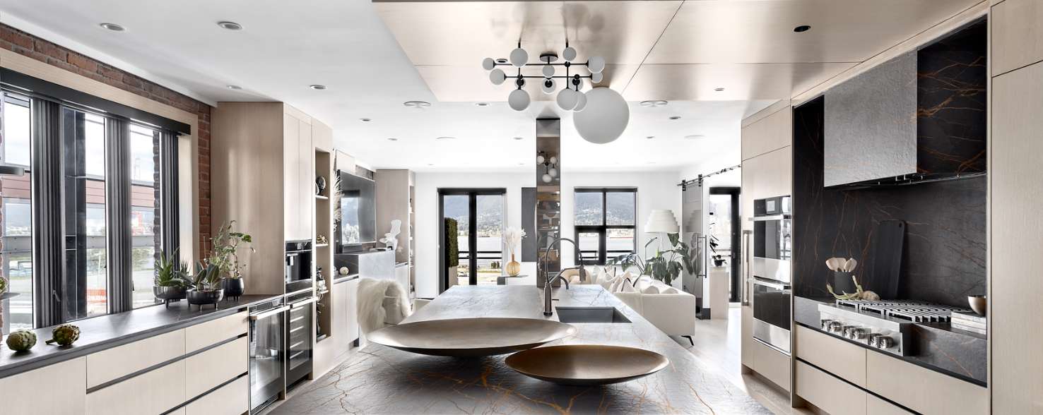 A kitchen with a long island under a fixture. On the left, there's floor to ceiling windows in front of a counter with potted plants on top. On the right is a JennAir Cooktop below an over-the-range hood. In the background is a family room with a white couch in front of a television, surrounded by shelves with various items on them. Behind the living room are a large potted plant, a standing lamp, windows and glass doors.