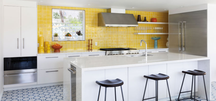 A kitchen with a yellow backsplash. There's a JennAir Range below an Over-the-Range Hood. On the counter are bowls and vases and a shelf of wine glasses. There's also a JennAir Microwave and an island with a sink and three chairs.