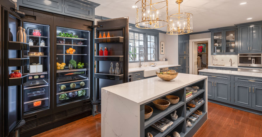 A kitchen with an opened JennAir Refrigerator. The refrigerator is filled with beverages, produce in crispers and bowls, fresh herbs, a cake box and more. There's a JennAir Microwave over a JennAir Wall Oven. Nearby is an island with shelves filled with bowls, a mortar and pestle and cookbooks.