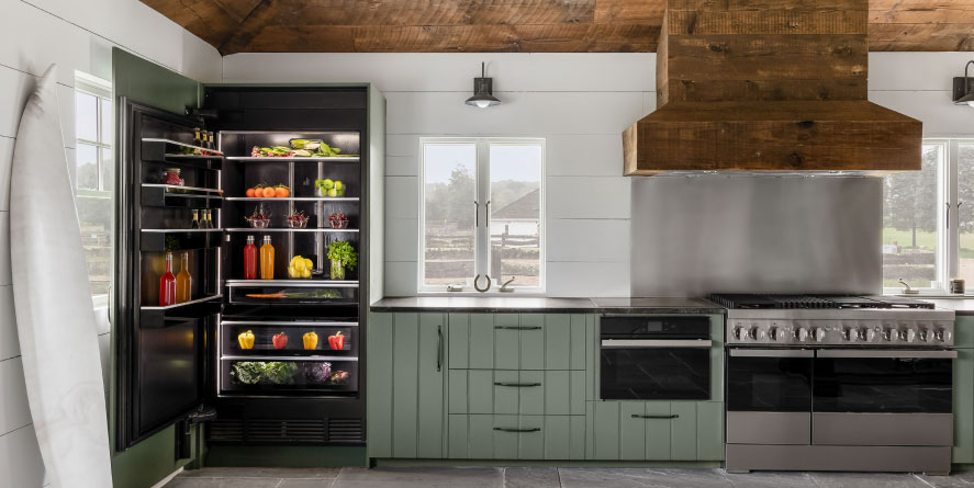 A kitchen with an opened JennAir Refrigerator. Inside the refrigerator are beverages, fresh herbs, produce and fruit bowls. Next to it are green cabinets, a JennAir Microwave and a JennAir Double Oven below an Over-the-Range Hood.