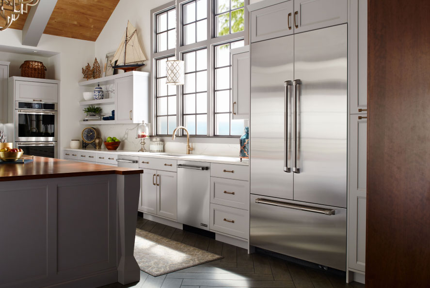 A kitchen with a JennAir French Door Refrigerator with a Bottom Freezer. On the counter next to it are a fruit bowl and various items. Next to the counter is a JennAir Double Wall Oven. In the foreground is an island with a fruit bowl on it.
