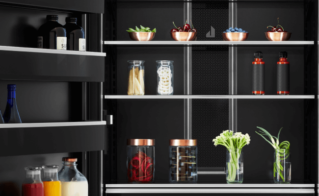 Réfrigérateur haut de gamme JennAir avec la porte ouverte, dévoilant divers articles comme des fines herbes dans des verres, des bols de cerises, de bleuets et d’edamames et des boissons.