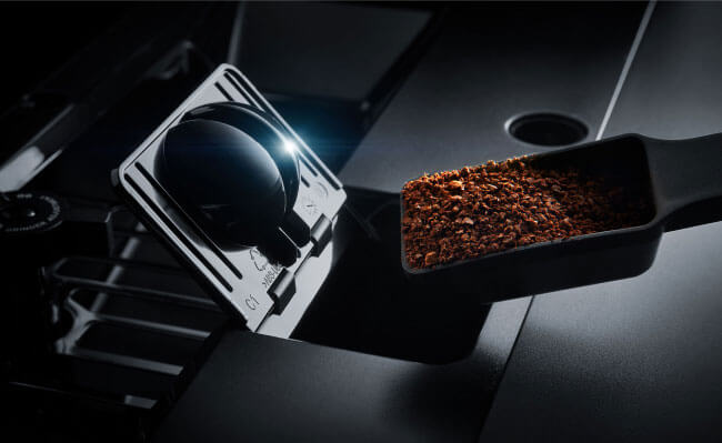 A scooper filled with coffee grounds over the compartment of a coffee maker where you enter the grounds.
