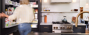 Une femme floue se tient devant un réfrigérateur JennAir® ouvert et rempli d’aliments et de boissons. À côté du réfrigérateur se trouve une cuisinière JennAir® avec une théière sur l’un des brûleurs.