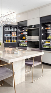 : A stunning kitchen with two JennAir fridges between a JennAir Double Wall Oven. The fridges are open and full of contents, like beverages and produce. In front is a marble dining room table with chairs. 