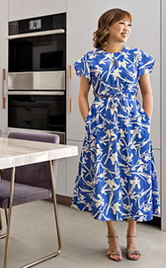 A flawless kitchen with a JennAir Double Wall Oven. A woman in a blue and white dress stares off, smiling, hands in pockets. She's next to a table with glasses, plates and a flower centrepiece. 