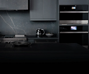 A modern high-end kitchen with a marble backsplash and black cabinets. There's a JennAir cooktop with a wok on a burner and JennAir double wall ovens. On the counter are a coffee carafe, a bowl and several containers filled with various items.