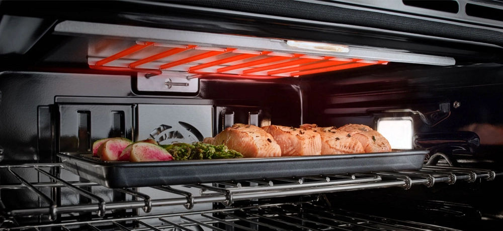 L’intérieur d’un four encastré JennAir avec l’élément chauffant allumé. Une plaque de cuisson contenant des légumes et des filets de saumon se trouve sur la grille du haut.